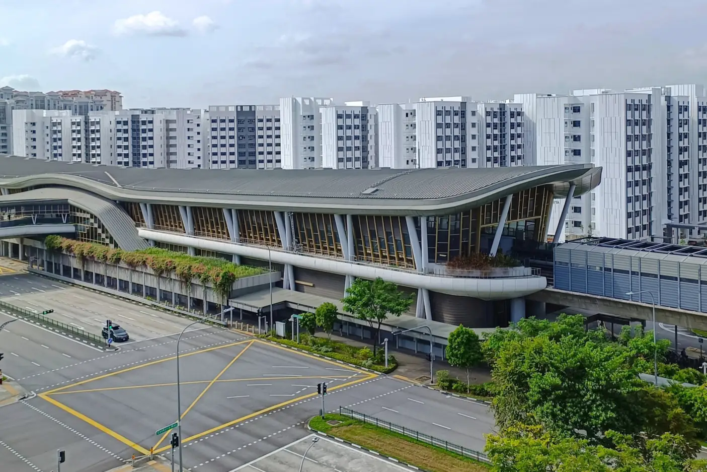 Elevated view of Canberra MRT station with its curved grey roof and timber-slatted facade, a quiet road junction in front and rows of white HDB blocks behind