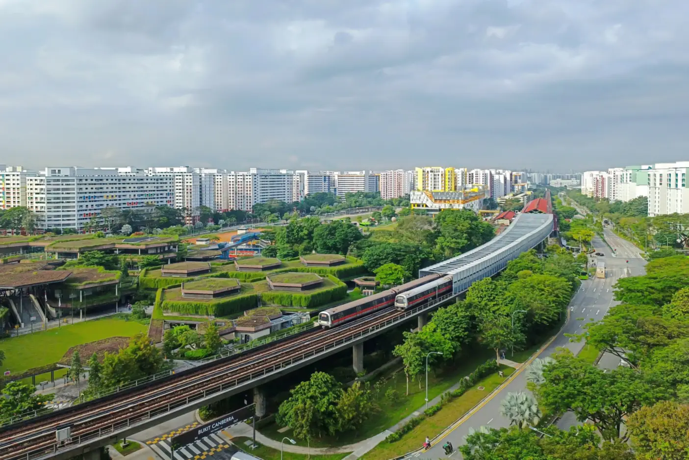 Aerial view over Bukit Canberra in Sembawang, with a North-South Line train on the elevated track, green-roofed sports buildings, tree-lined roads and HDB blocks stretching to the horizon