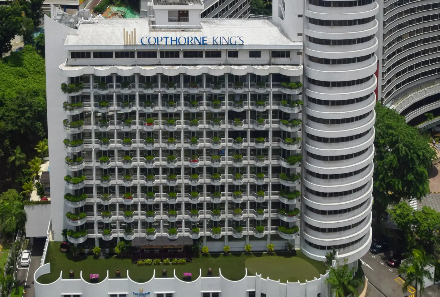Aerial view of the white Copthorne King's Hotel on Havelock Road, its rows of curved balconies lined with potted plants, a rounded tower beside it and a rooftop lawn over the podium