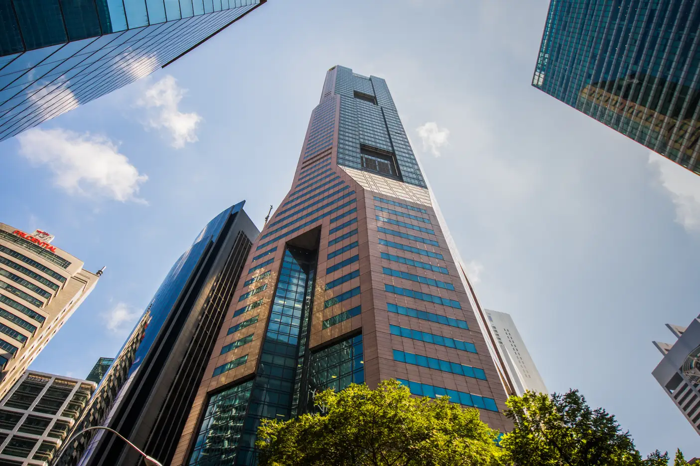 Looking up at Republic Plaza, a tall bronze-clad office tower with blue glass bands, framed by neighbouring Raffles Place towers, trees and a blue sky with light clouds