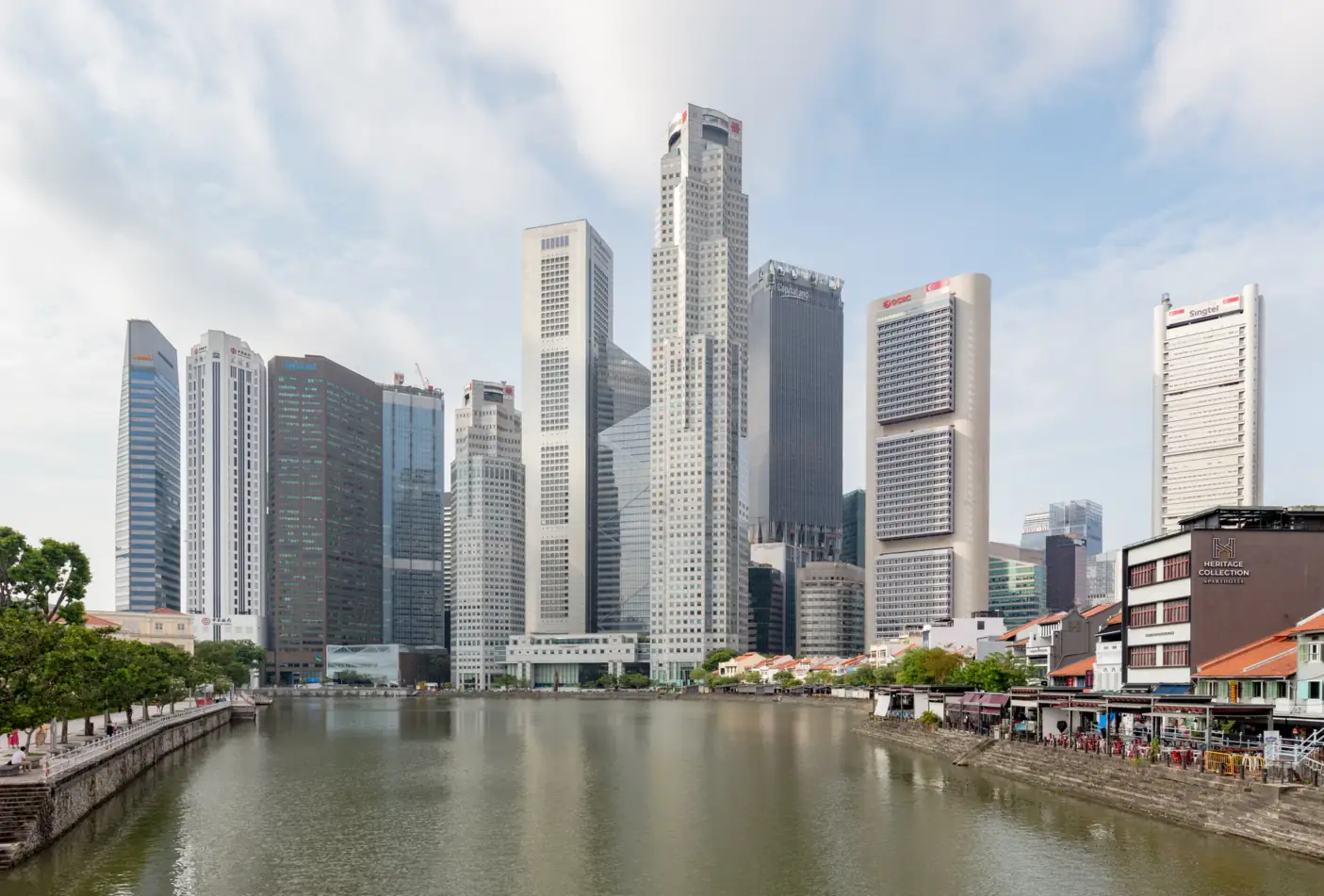 Office towers of Singapore's central business district rising behind the Singapore River, with low-rise conserved shophouses and riverside dining along the right bank under a pale blue sky