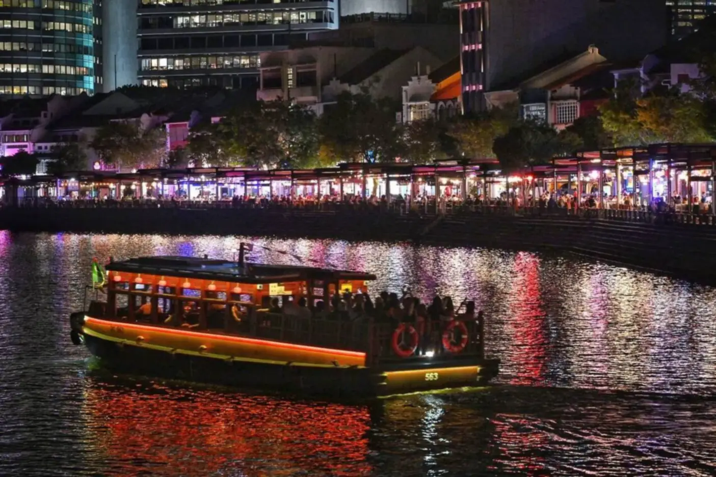 A lit bumboat crossing the Singapore River at night in front of the Boat Quay waterfront, where rows of outdoor restaurant and bar seating glow beneath conserved shophouses and office towers