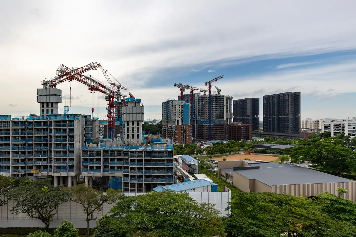 Residential blocks under construction wrapped in blue safety netting, with red tower cranes overhead, a hoarded site and trees in front, and taller dark residential towers in the background