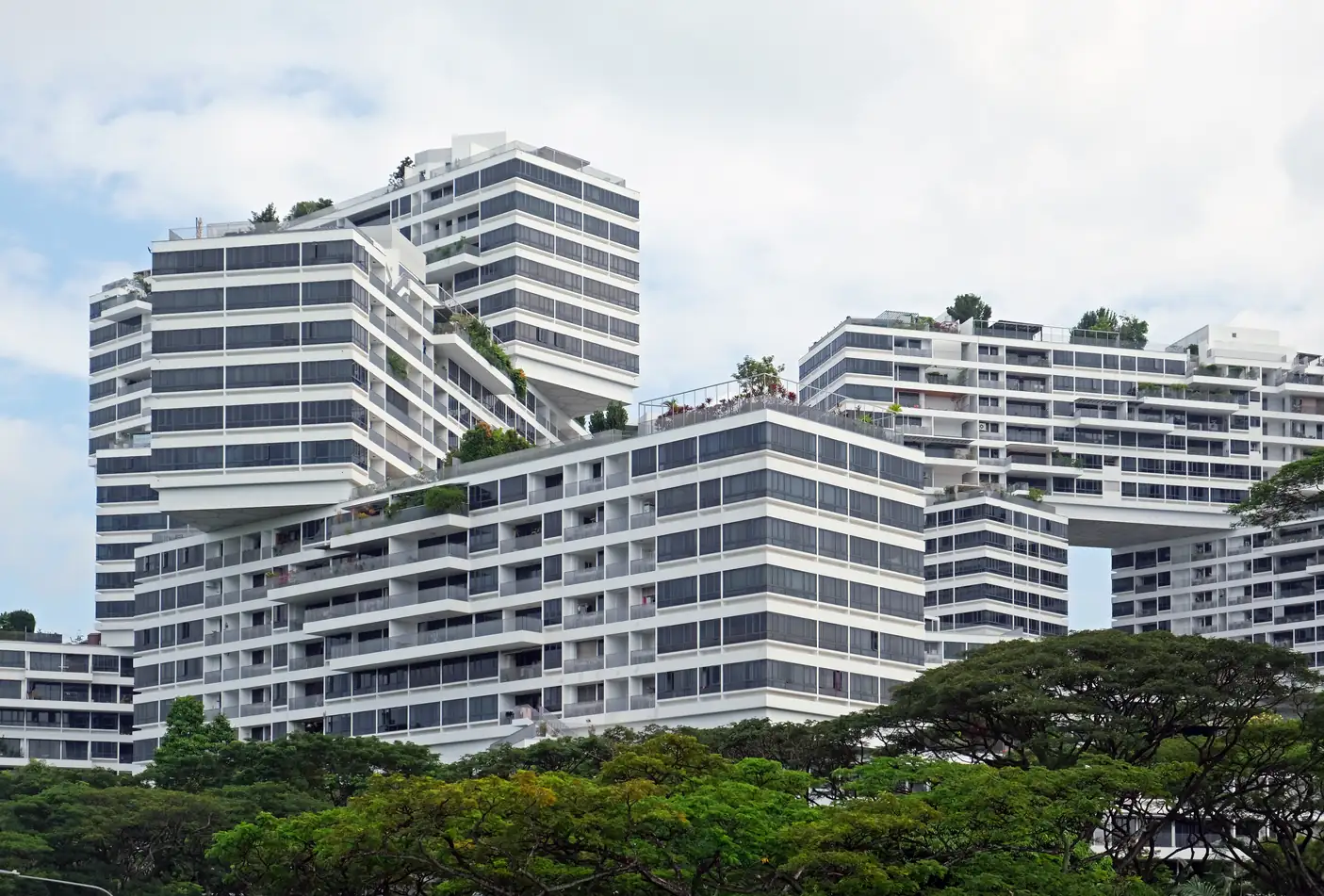 The Interlace apartment complex in Singapore, white residential blocks stacked at angles with planted roof terraces and deep balconies, rising above a canopy of mature trees under a cloudy sky