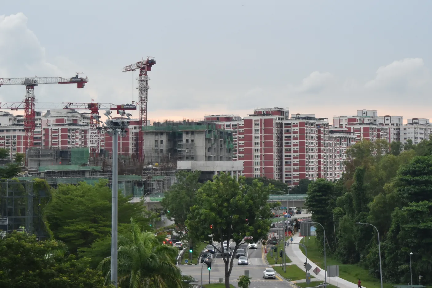 Tower cranes over HDB blocks still under construction, beside completed red-and-white HDB blocks, with a tree-lined road and traffic junction in the foreground