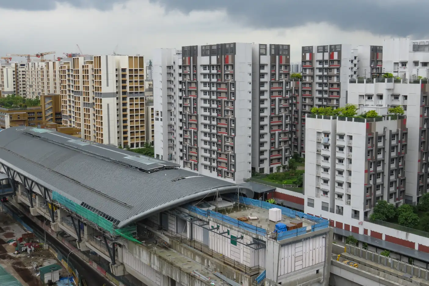 Elevated view of Tengah Plantation MRT station under construction, its curved grey roof beside new white HDB blocks with red and grey facades and rooftop gardens