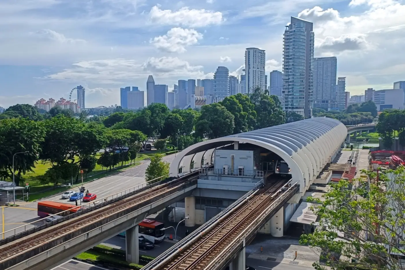 The curved, ribbed roof of Kallang MRT station over elevated East-West Line tracks, with a train at the platform, trees and a road junction in front and the city skyline behind