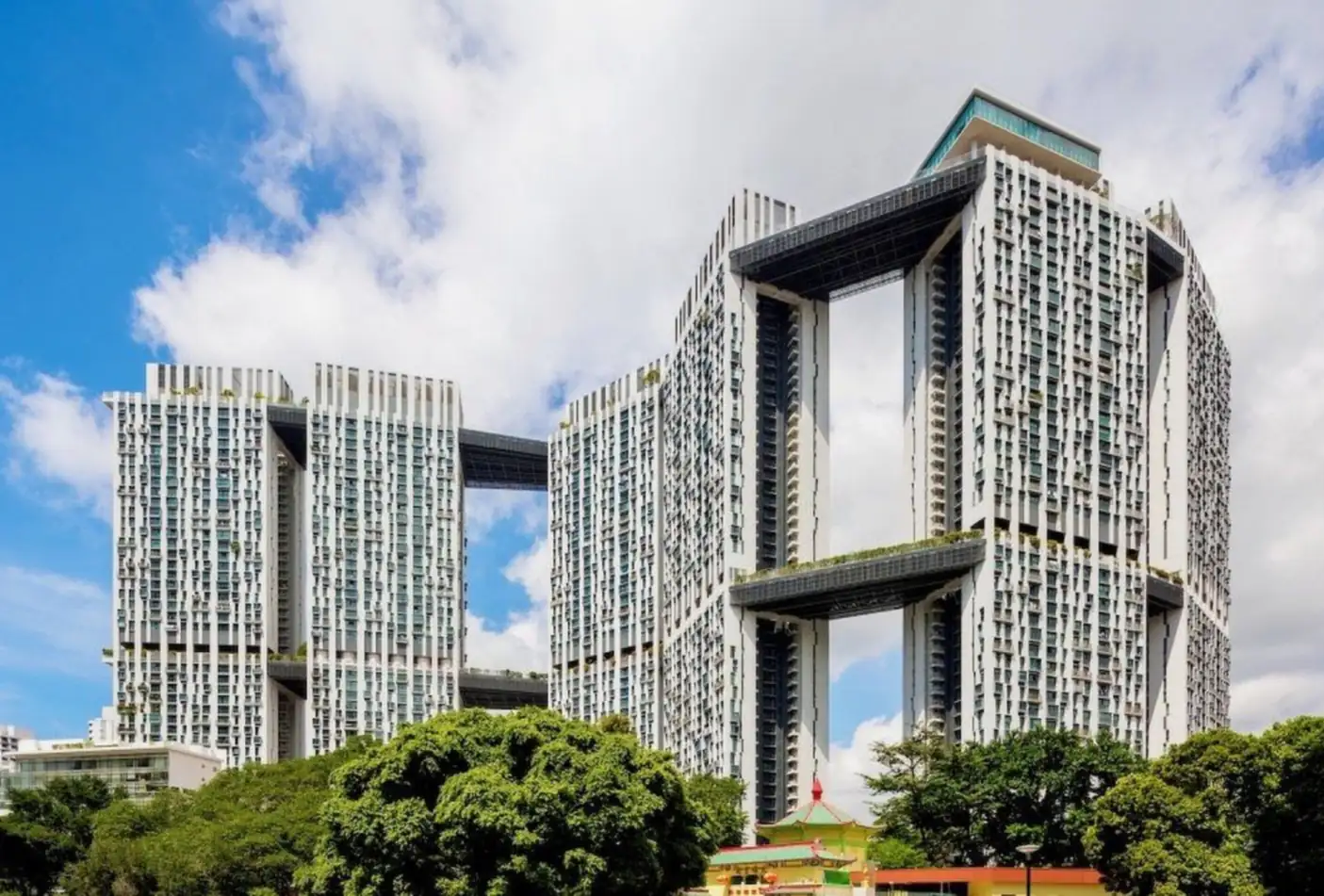 The Pinnacle@Duxton, the 50-storey public housing development in Tanjong Pagar, its tower blocks linked by sky bridges, seen from street level against a bright sky with trees and a red-and-green Chinese-style roof in front
