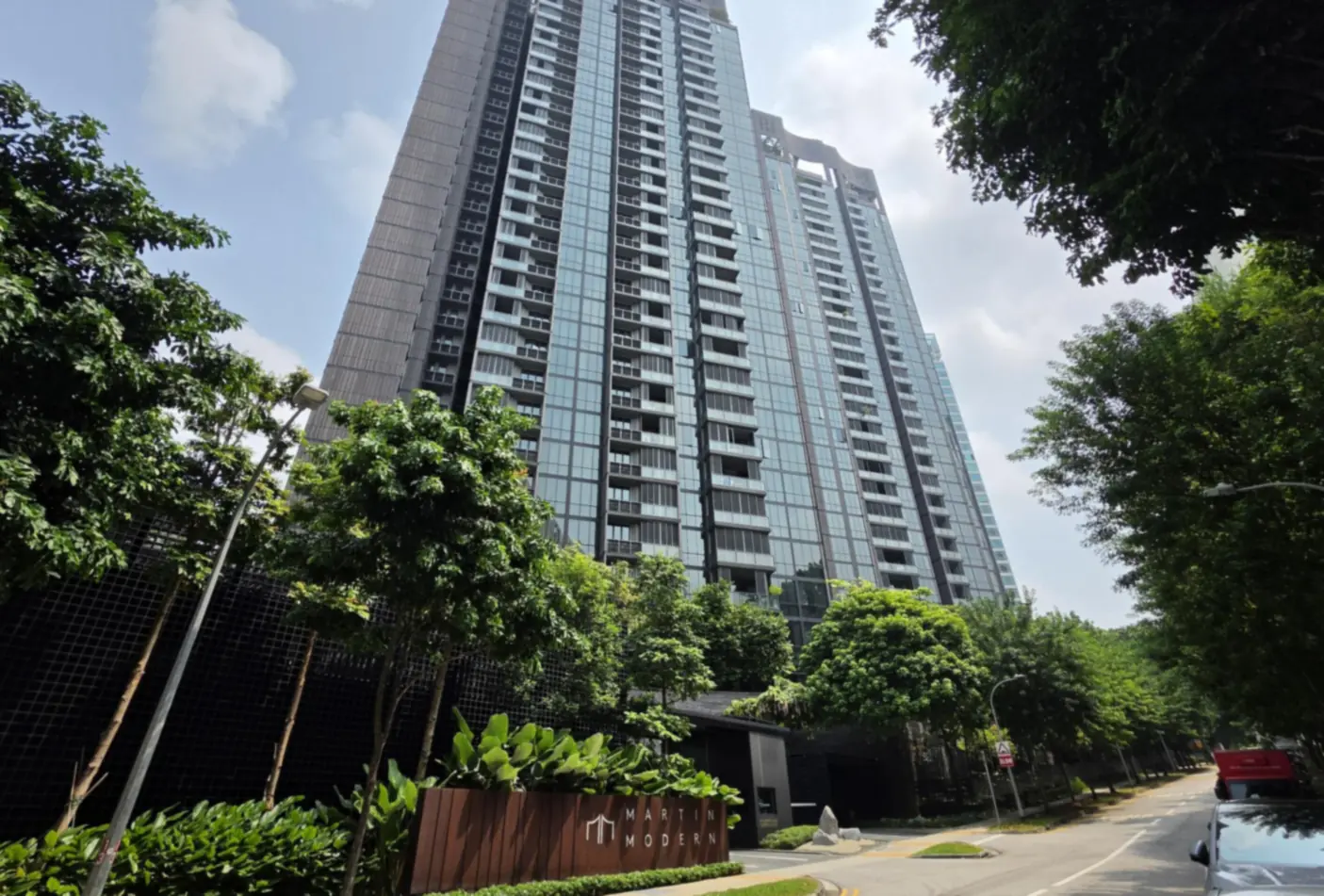 Martin Modern condominium towers in River Valley seen from street level behind leafy trees, with the development's timber entrance sign in the foreground and a quiet road on the right