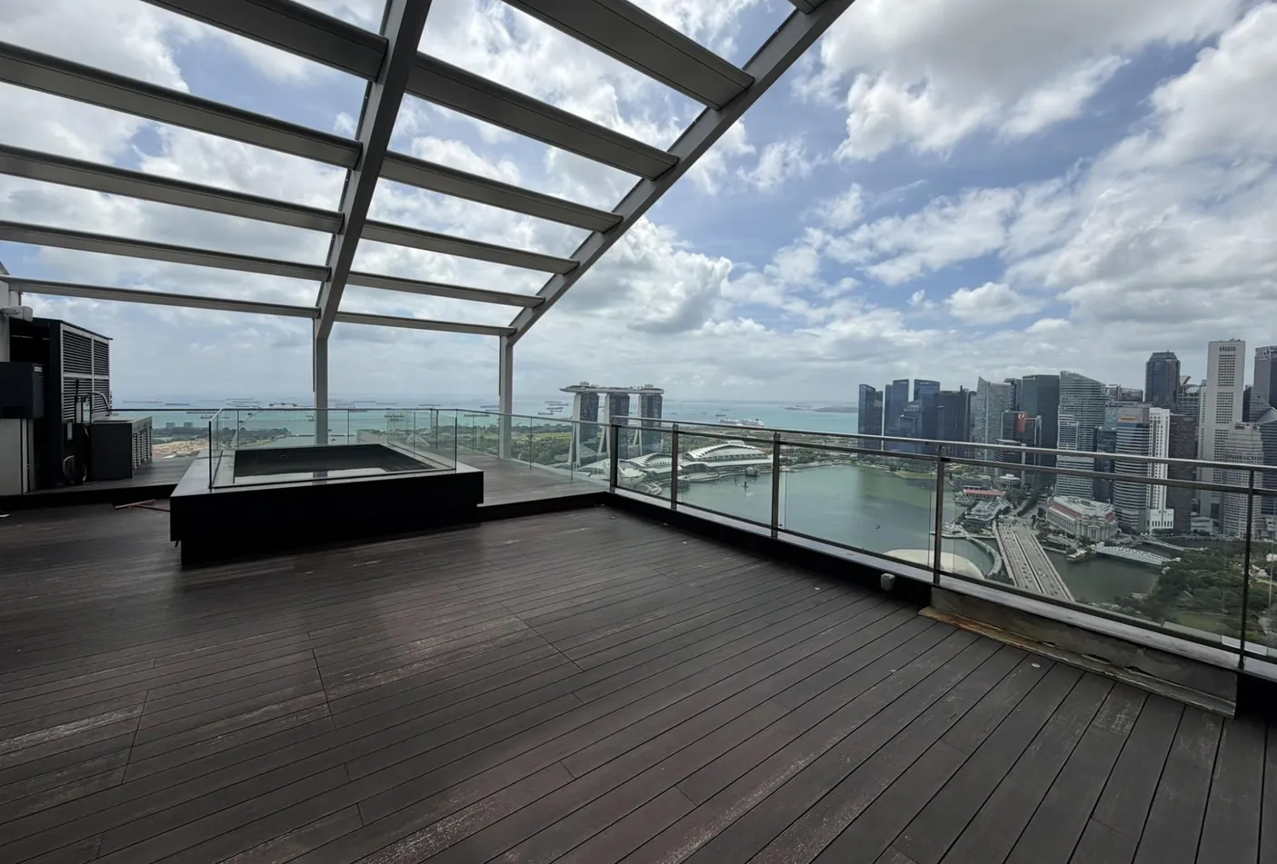 Empty roof terrace of the South Beach Residences penthouse, with timber decking, a raised pool and glass railings overlooking Marina Bay Sands, the bay and the Singapore CBD skyline