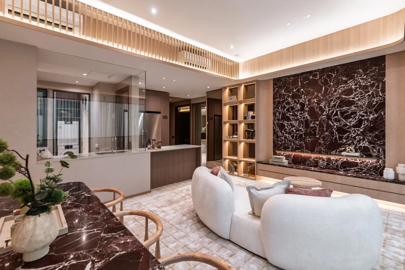 Living and dining area of a four-bedroom showflat, with a curved white sofa, a red marble feature wall and dining table, lit timber shelving, slatted timber ceiling detail and an open kitchen behind glass