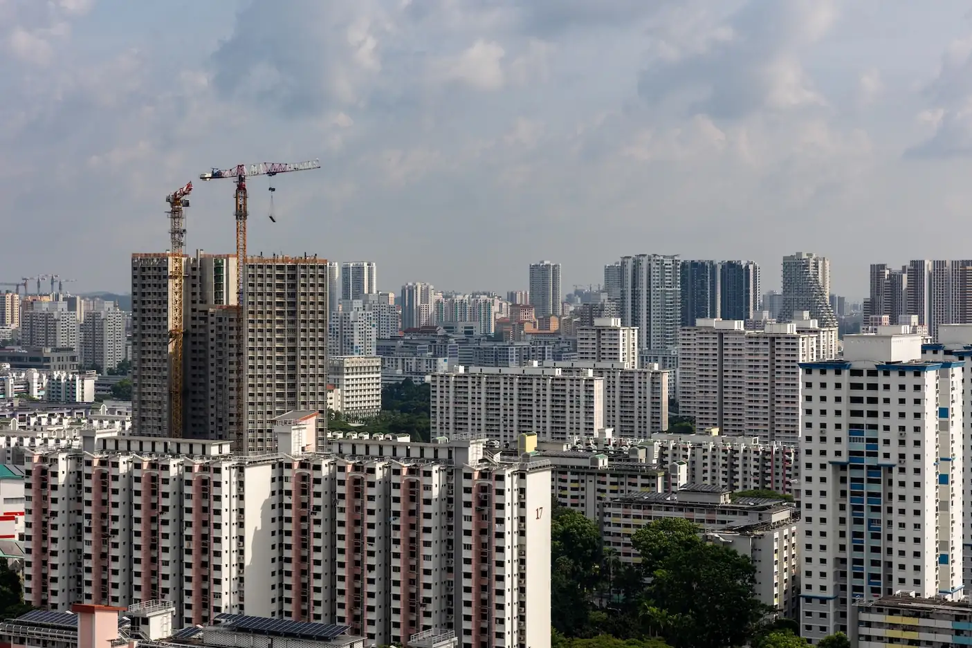 Rows of HDB blocks in a mature estate seen from above, with a high-rise residential block under construction beneath two tower cranes and more residential towers stretching to the horizon under a cloudy sky