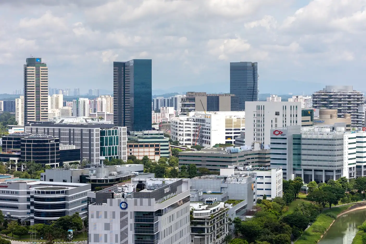 Elevated view of the International Business Park, with glass office towers, low-rise business park blocks and rooftop gardens set among trees beside a canal, and HDB estates on the horizon under a cloudy sky