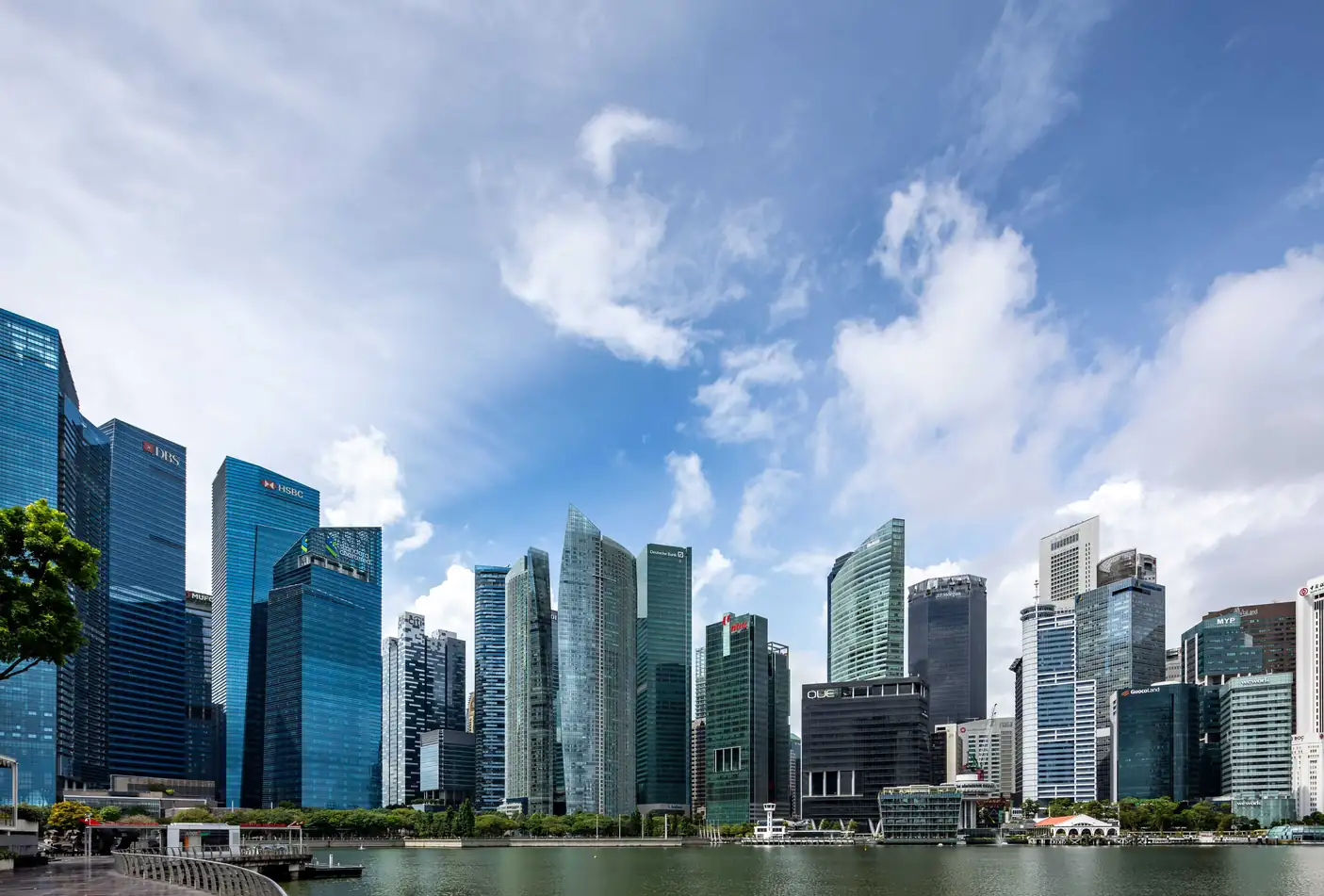 Singapore's CBD office towers rising above the Marina Bay waterfront under a blue sky with scattered white clouds, with a tree and an empty promenade in the foreground