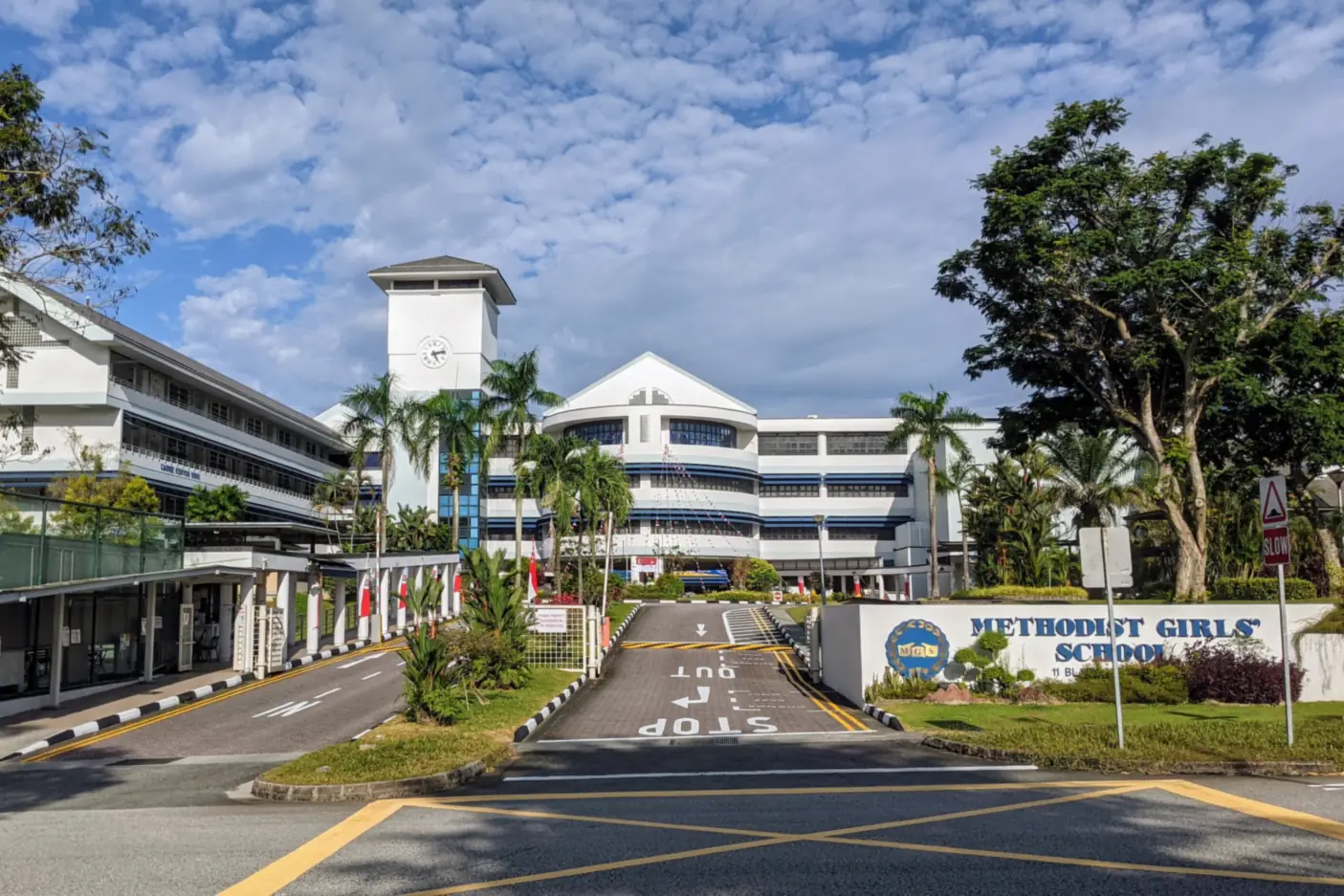 Methodist Girls' School at Blackmore Drive: a white clock tower and blue-trimmed classroom blocks behind palm trees, with the school name on a low wall beside the entrance driveway