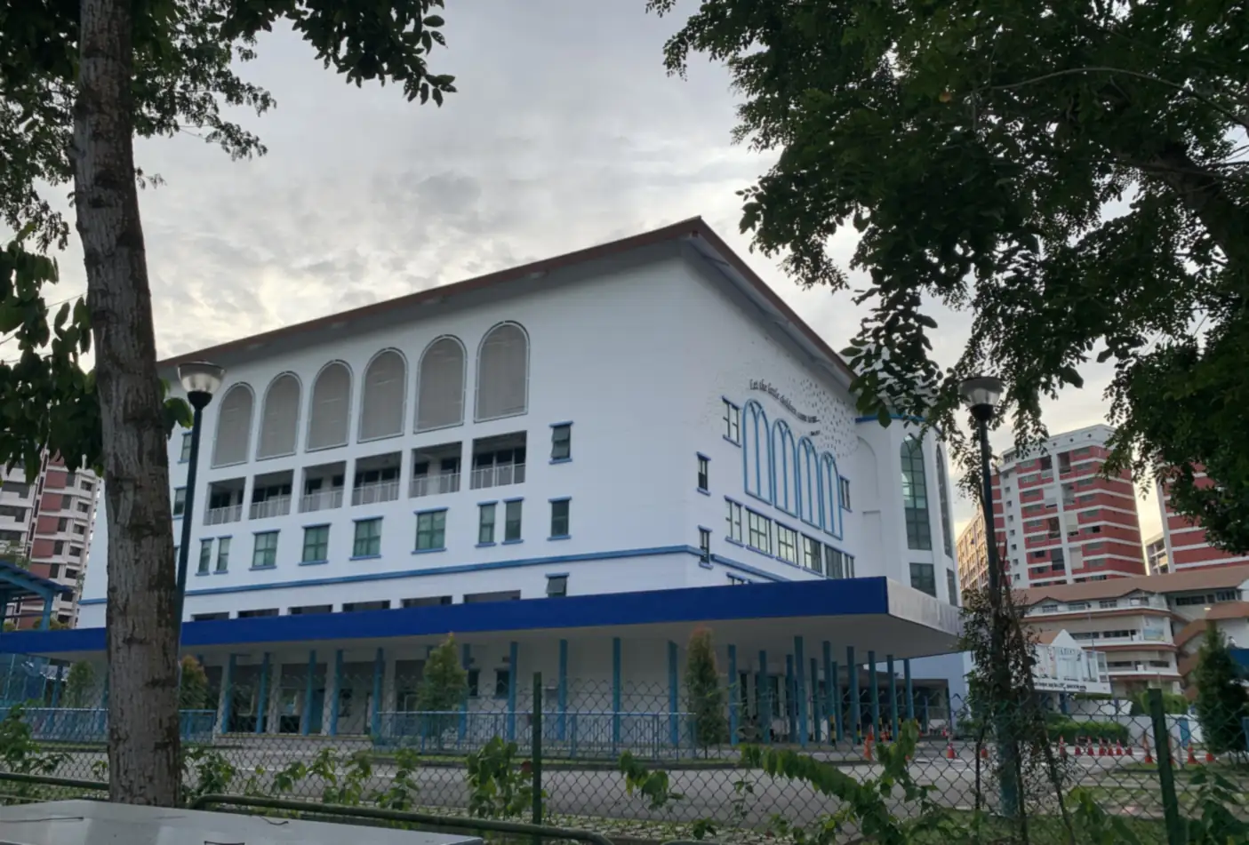 Pei Hwa Presbyterian Primary School, a white building with arched upper-floor windows and a blue band along its base, seen through trees and a chain-link fence, with residential blocks behind it