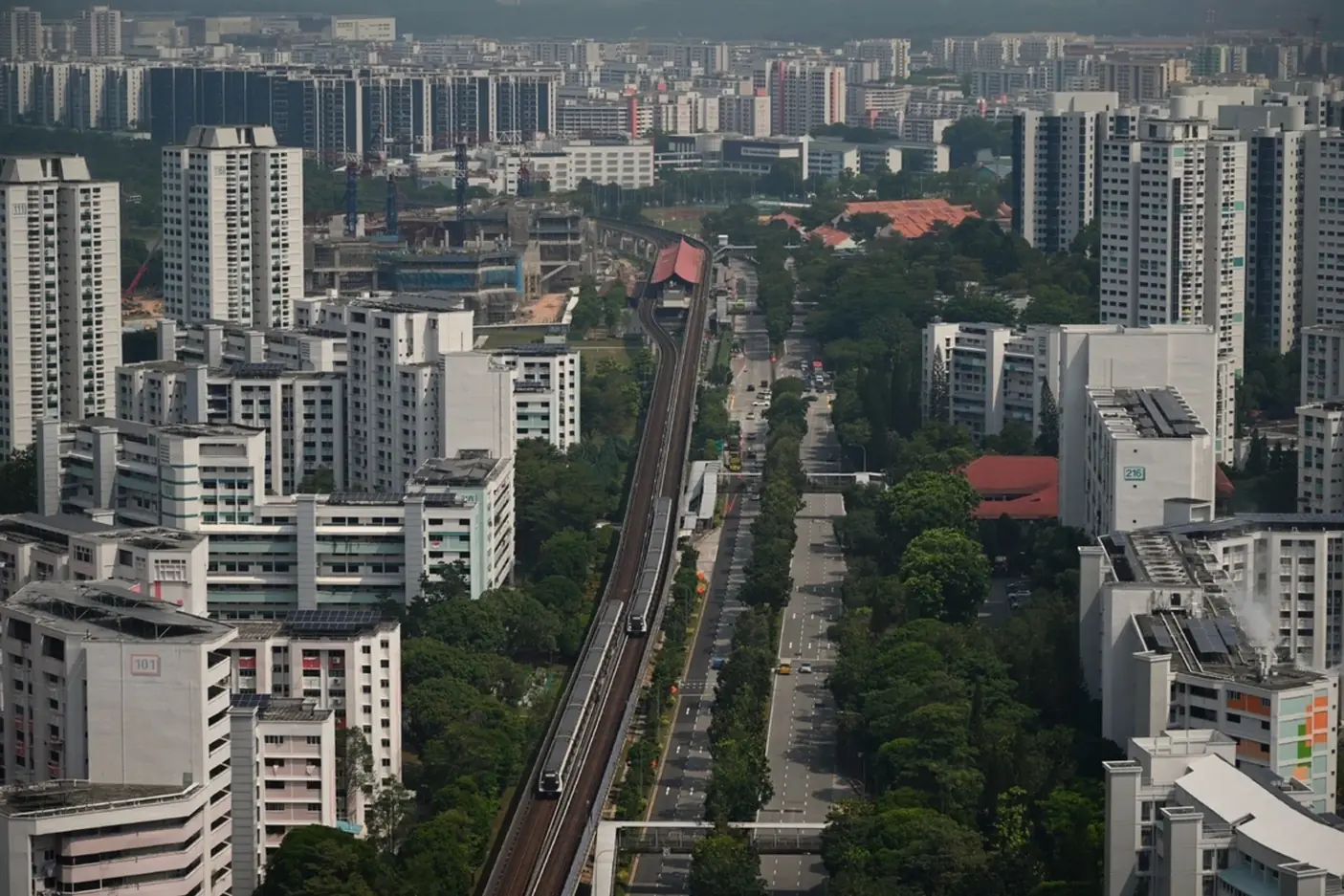 Aerial view of an elevated MRT line running beside a tree-lined road through an estate of high-rise housing blocks, with two trains on the viaduct and a station and construction site behind