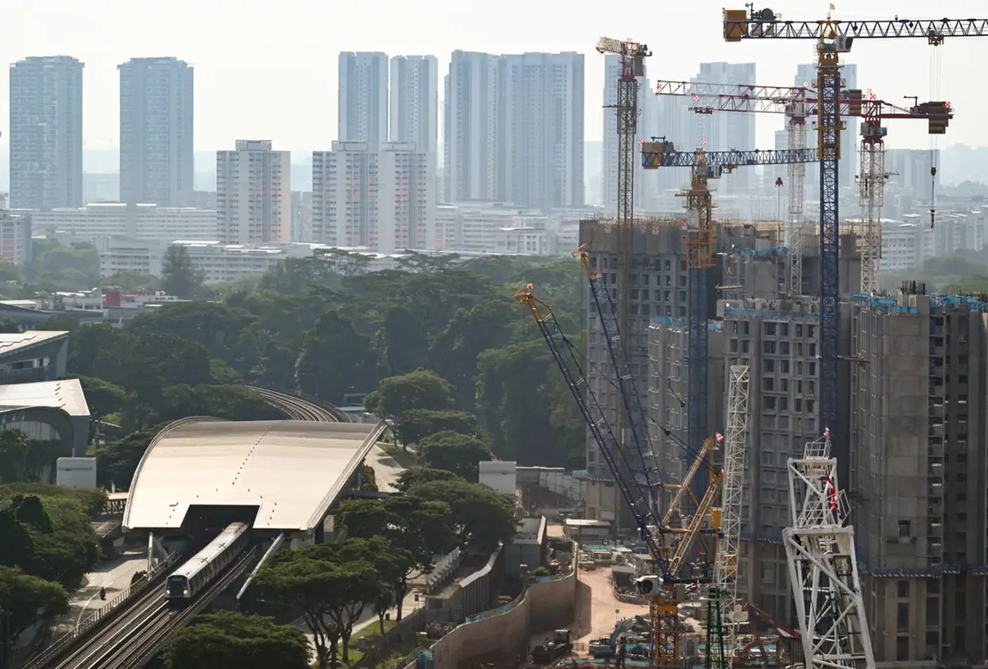 An MRT train on an elevated track pulls into an above-ground station beside high-rise blocks under construction, ringed by cranes, with high-rise housing behind