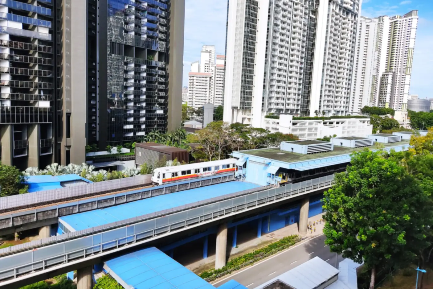 A train pulls into the elevated Queenstown MRT station, with a dark glass residential tower on the left and tall white apartment blocks behind, above a quiet road and trees