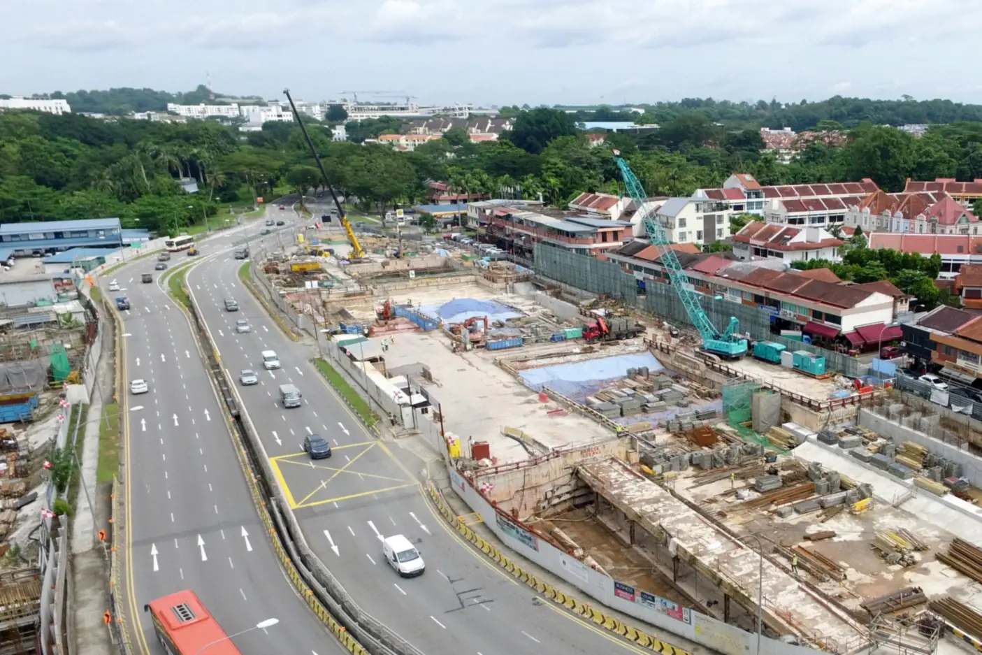 Aerial view of a large MRT station construction site beside a multi-lane road, with cranes and excavation next to a row of shophouses and red-roofed low-rise homes, and greenery beyond