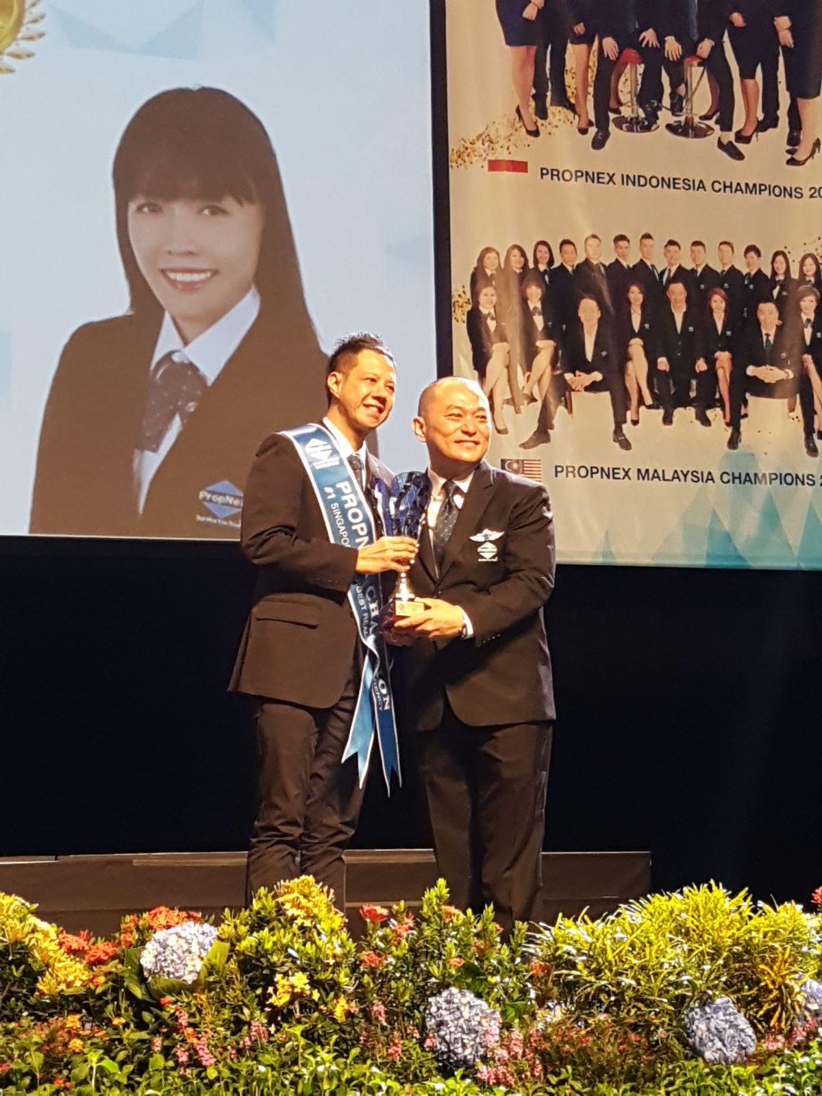Adrian on stage receiving a trophy from a PropNex leader at an Annual Convention, with Indonesia and Malaysia Champions backdrop