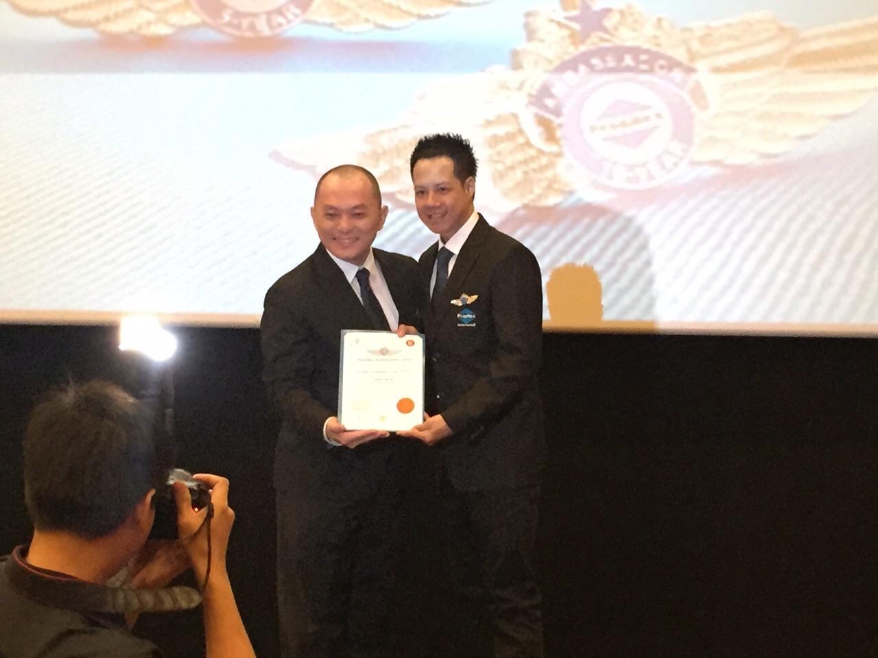 Adrian receiving a PropNex Ambassador certificate from a senior leader, with the 10-Year Ambassador badge projected on the screen behind them